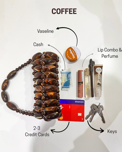 Collection of everyday items including a beaded bracelet, coffee, cash, lip combo, perfume, keys, and credit cards on a white surface, showing what can fit in the coffee handmade beaded bag