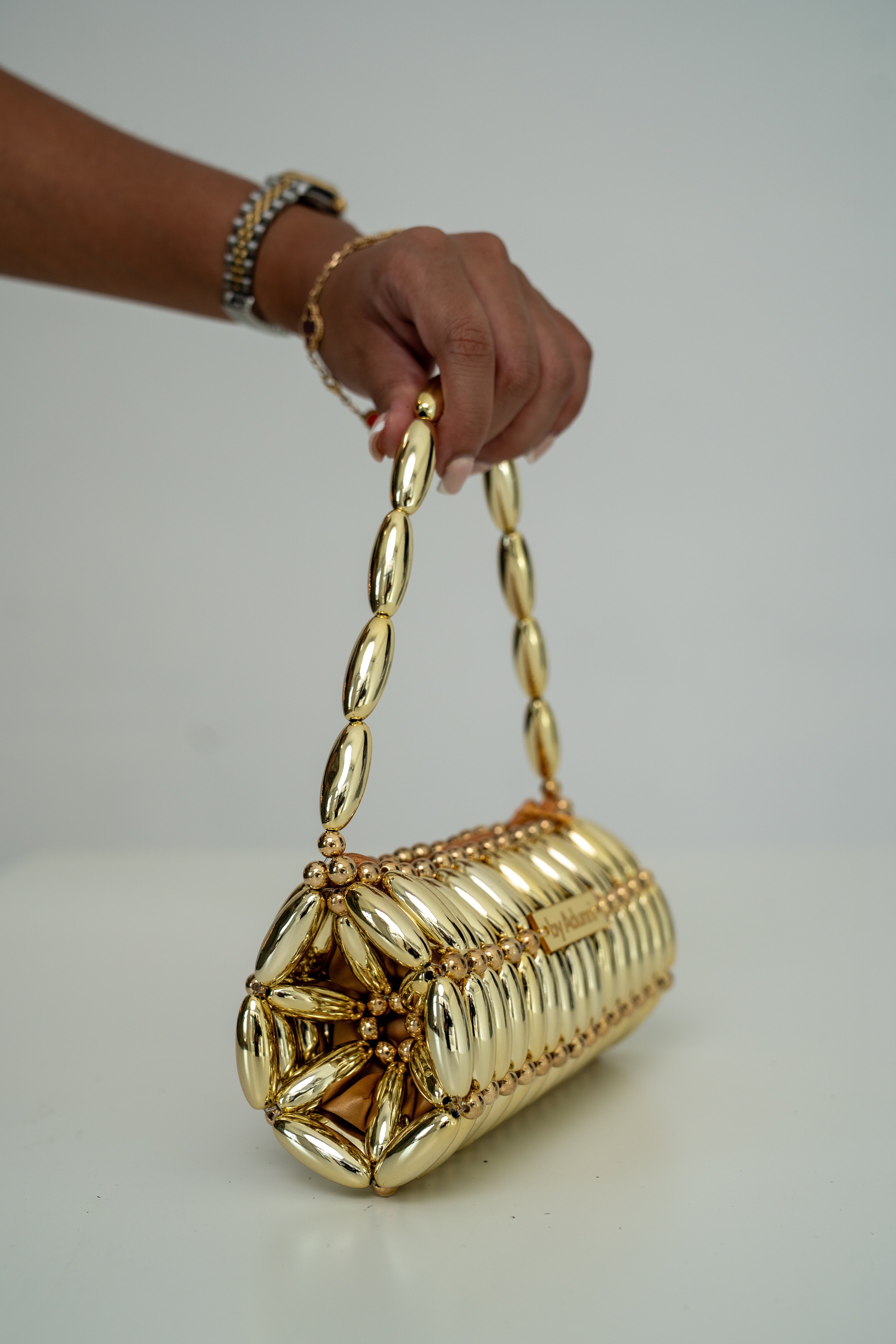 A hand holding a metallic gold cylindrical shoulder bag with a chain strap and a star-shaped embellishment on the front, set against a plain light gray background.