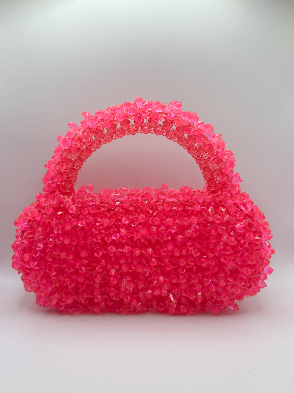 A small structured handbag covered in bright hot pink rhinestones, featuring a rounded body and a curved top handle, set against a neutral light gray background.