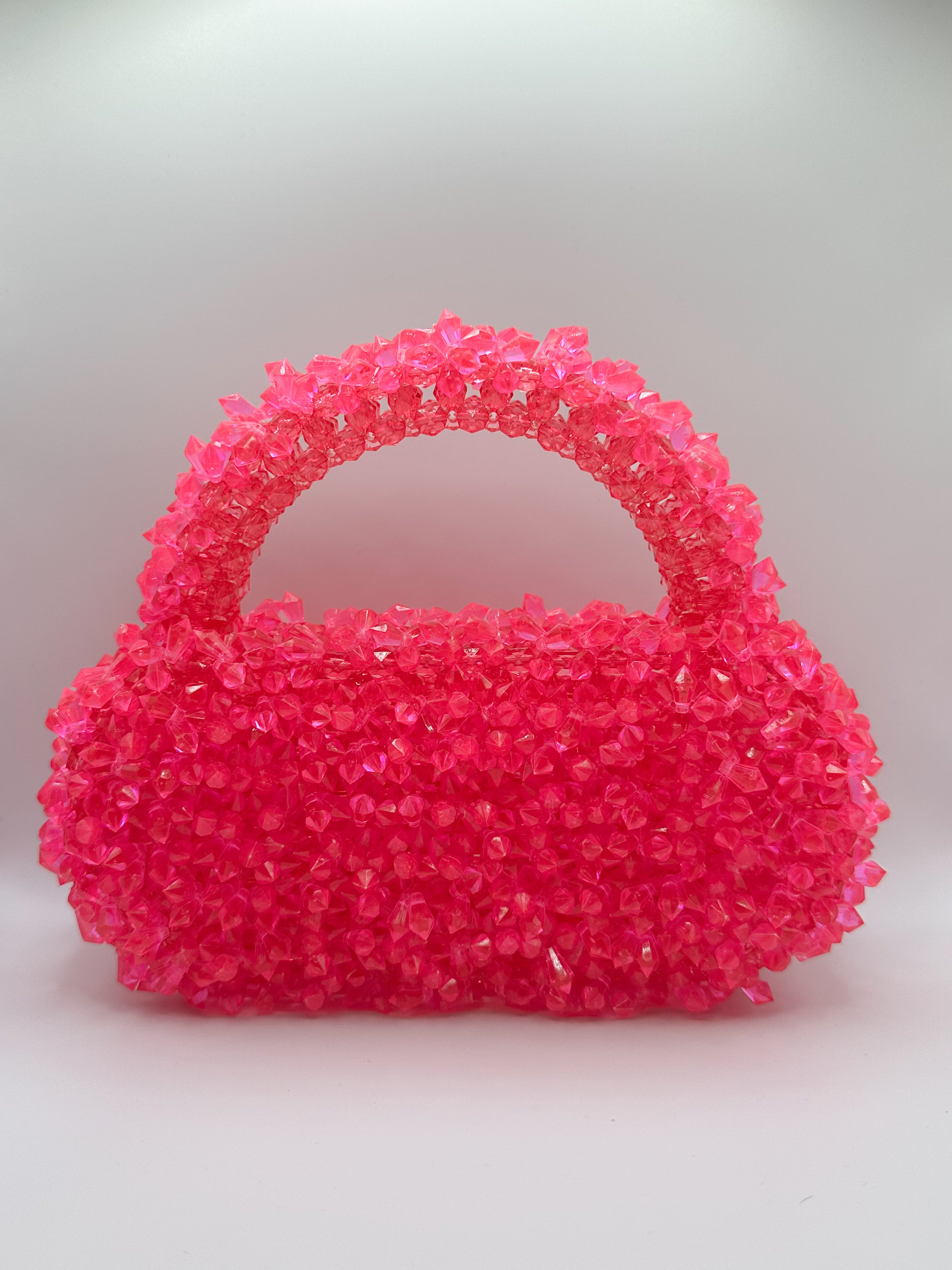 A small structured handbag covered in bright hot pink rhinestones, featuring a rounded body and a curved top handle, set against a neutral light gray background.