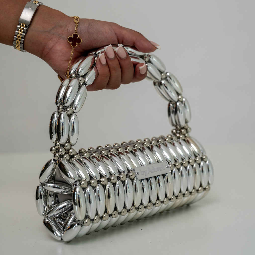 A silver metallic handbag with a unique textured, segmented body and a matching curved handle, held by a hand wearing gold bracelets against a plain light gray background.