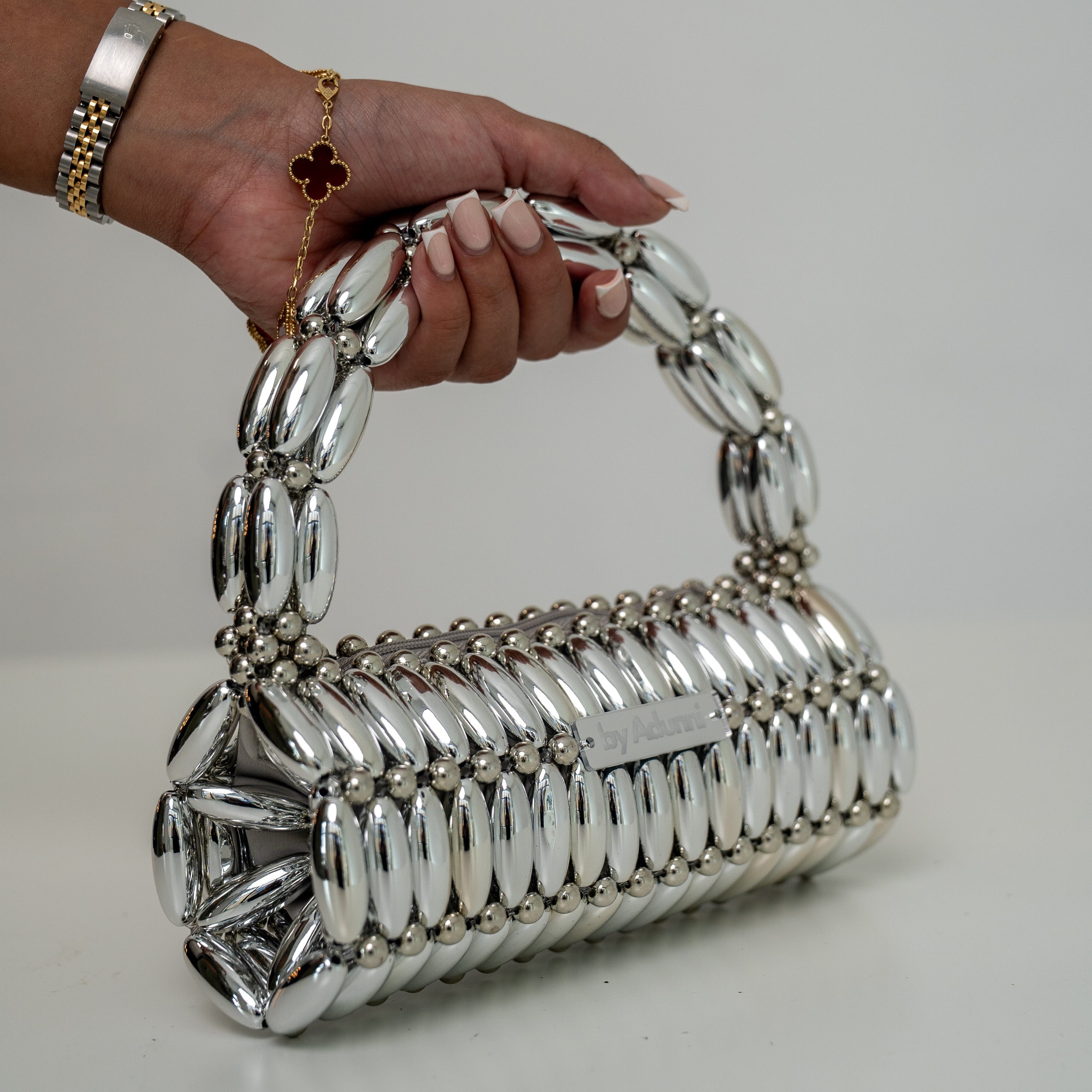 A silver metallic handbag with a unique textured, segmented body and a matching curved handle, held by a hand wearing gold bracelets against a plain light gray background.