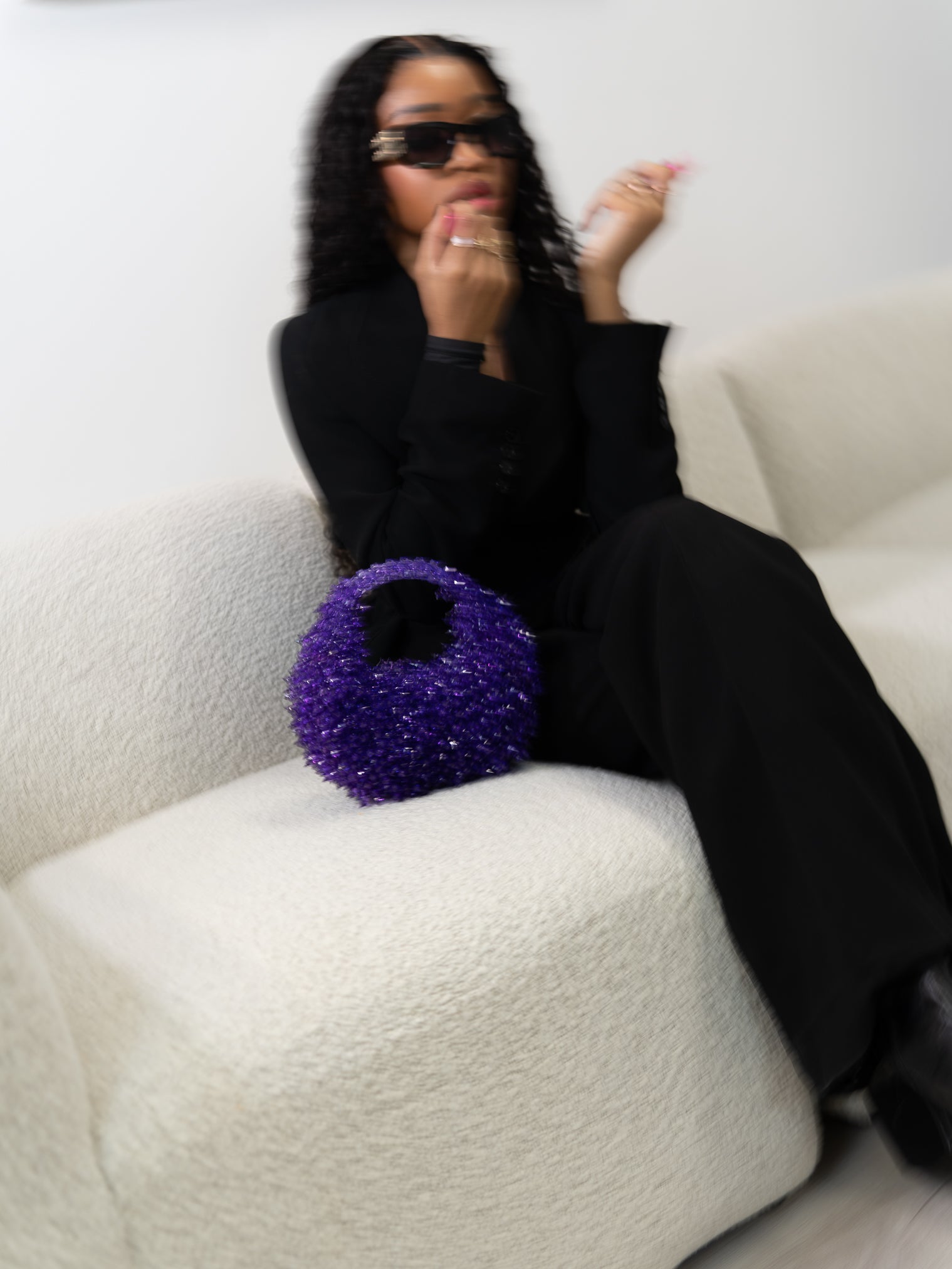 Person in black outfit with sunglasses holding a purple glittery bag on a white couch.