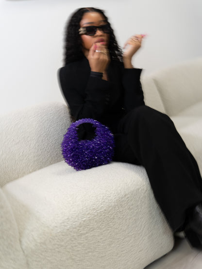 Person in black outfit with sunglasses holding a purple glittery bag on a white couch.