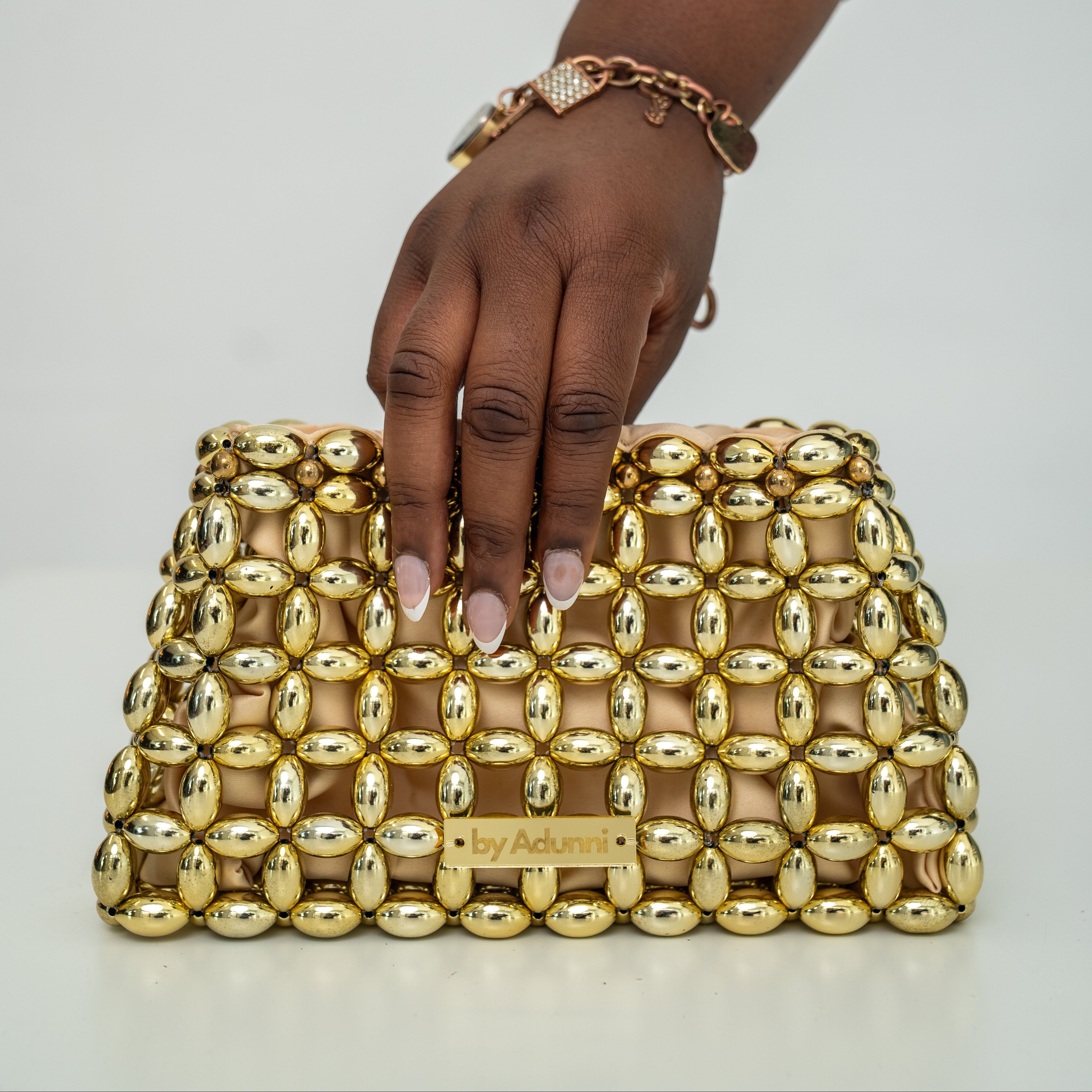 A gold beaded clutch bag with a small gold brand plaque on the front, held by a person's hand against a plain light gray background.