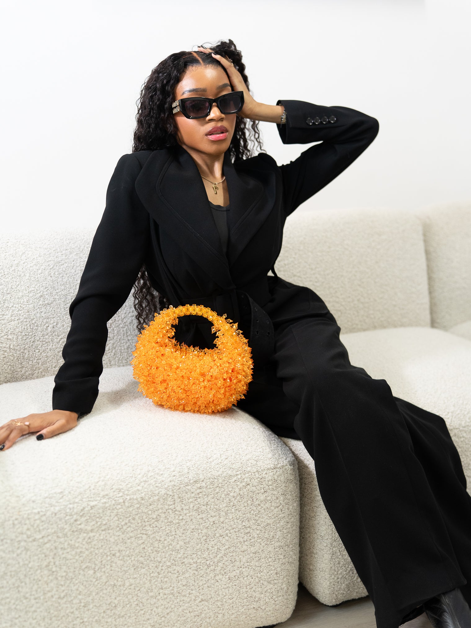 Woman in a black outfit with sunglasses holding an orange bag on a white couch.