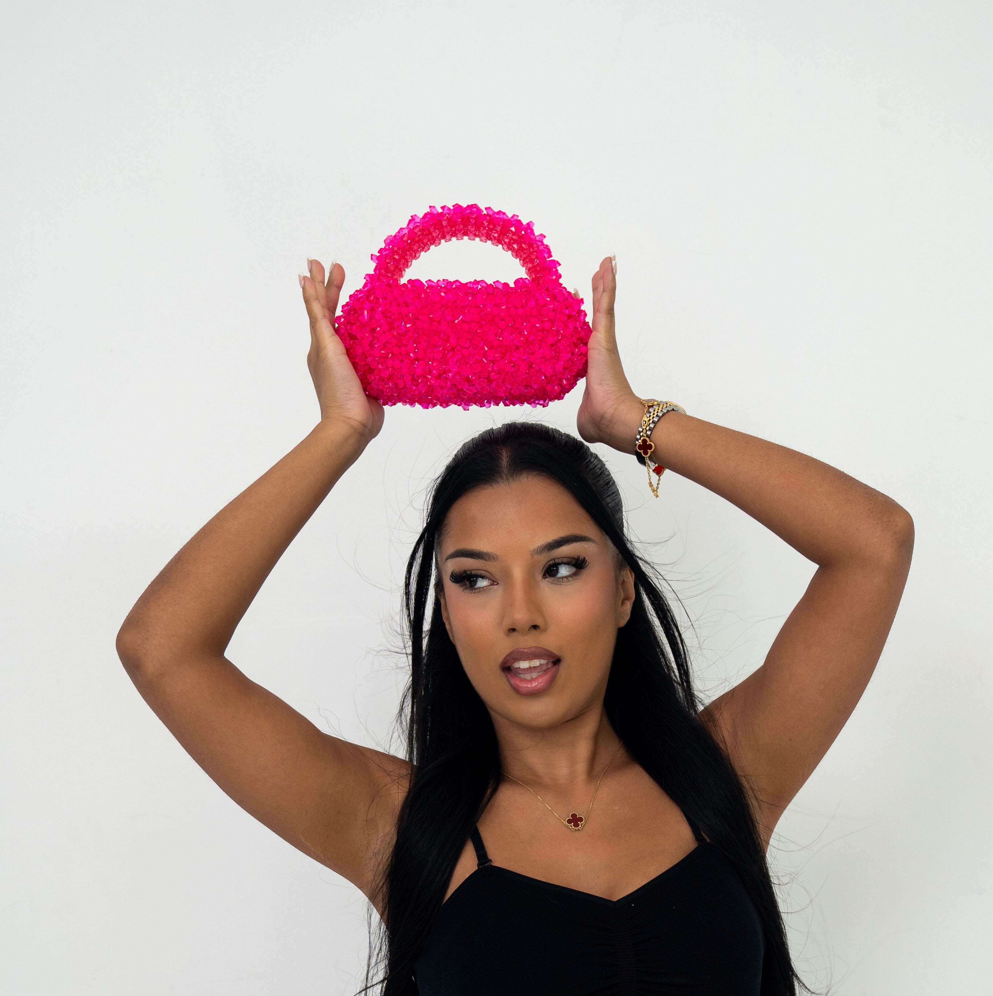 A woman with long black hair holds a bright hot pink textured handbag above her head with both hands, wearing a black top and gold jewelry against a plain white background.