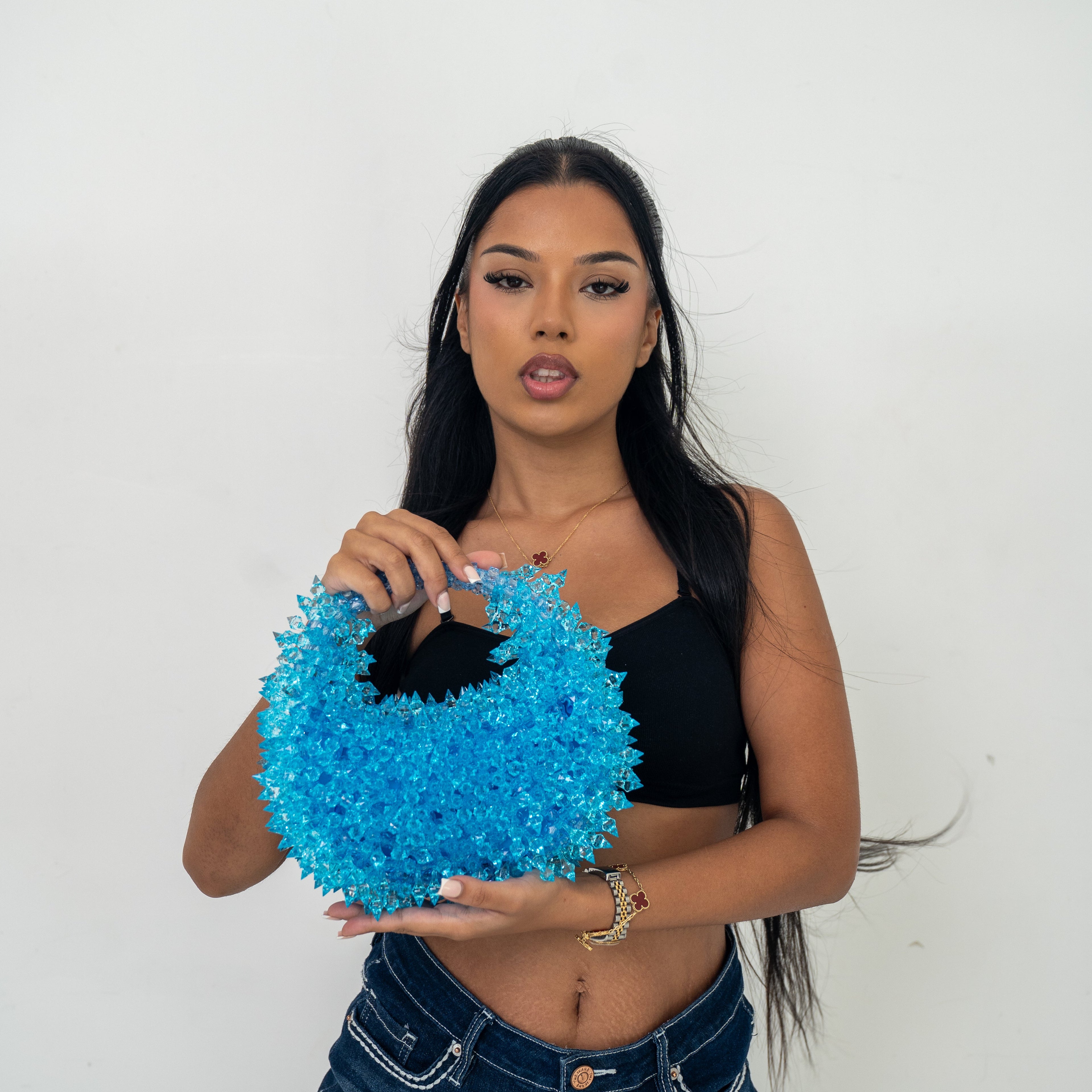 A woman modeling a bright blue, fuzzy, textured handbag with a black handle, paired with a black crop top and denim shorts, against a plain white background.