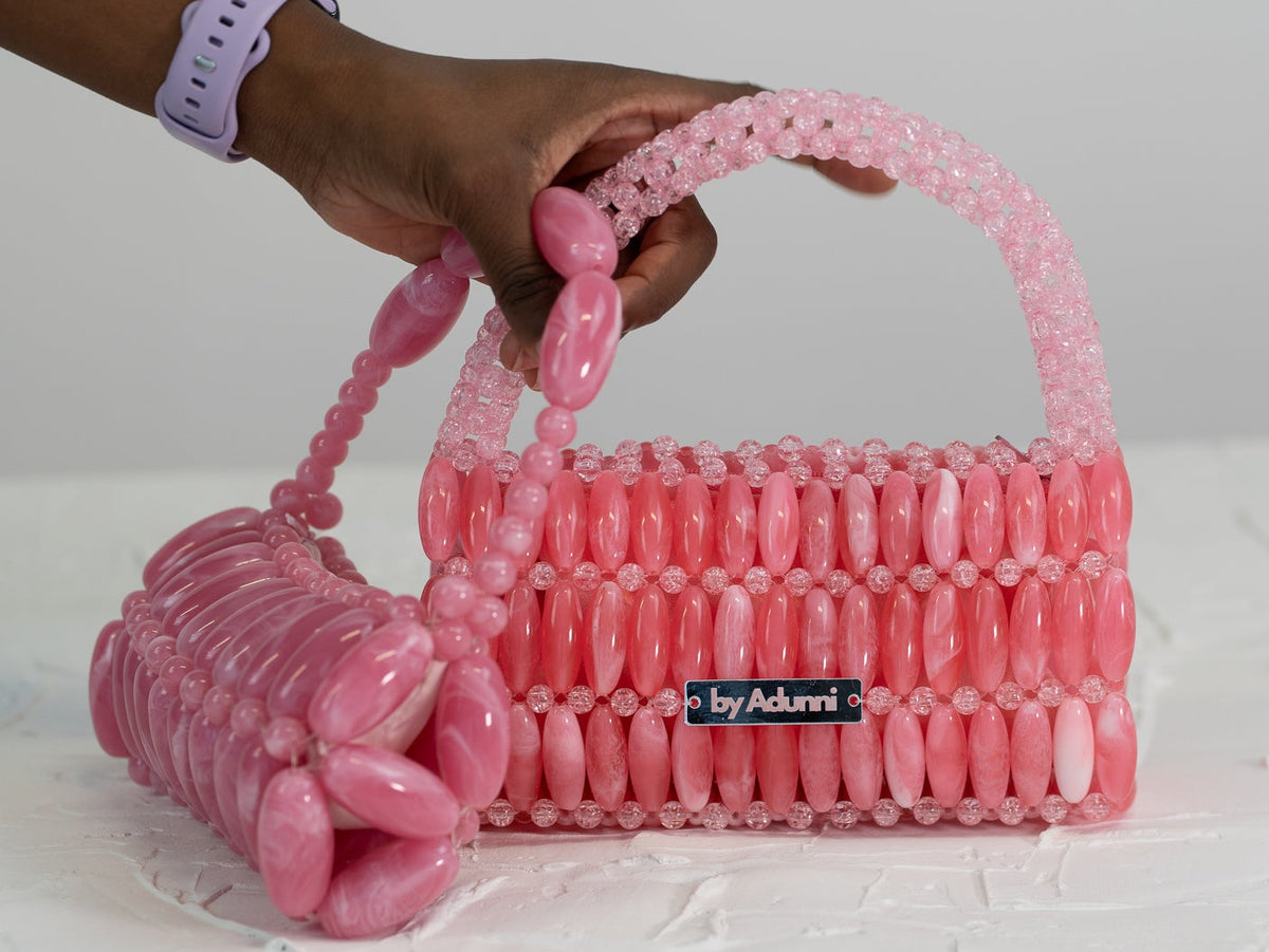 A hand holds a pink beaded handbag with a matching beaded wristlet strap against a light gray background. The bag has a small black label reading 'by Adunni'.
