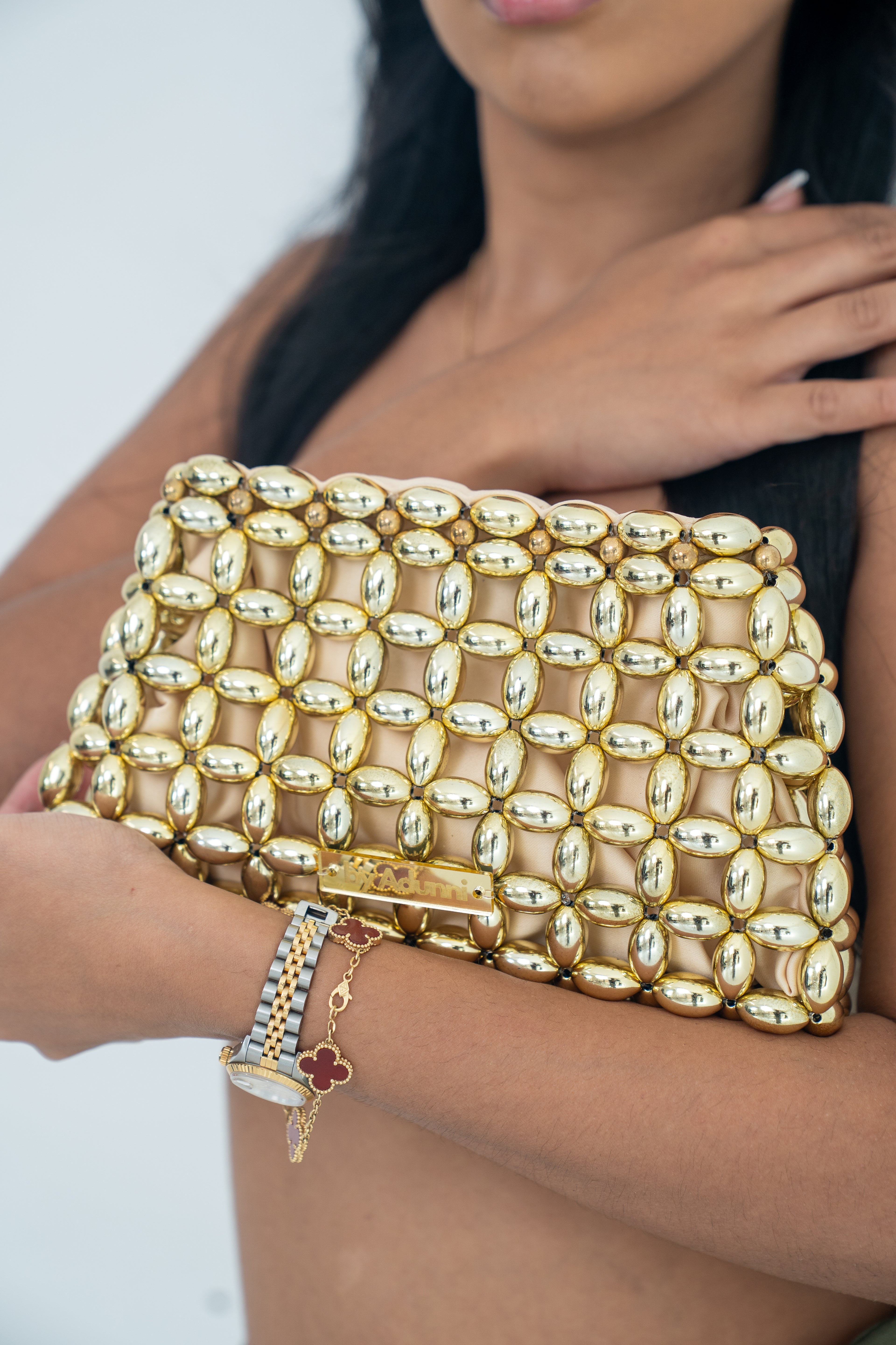 A close-up of a woman holding a structured gold clutch bag covered in a metallic mesh pattern of oval and round studs, with a gold clasp closure.
