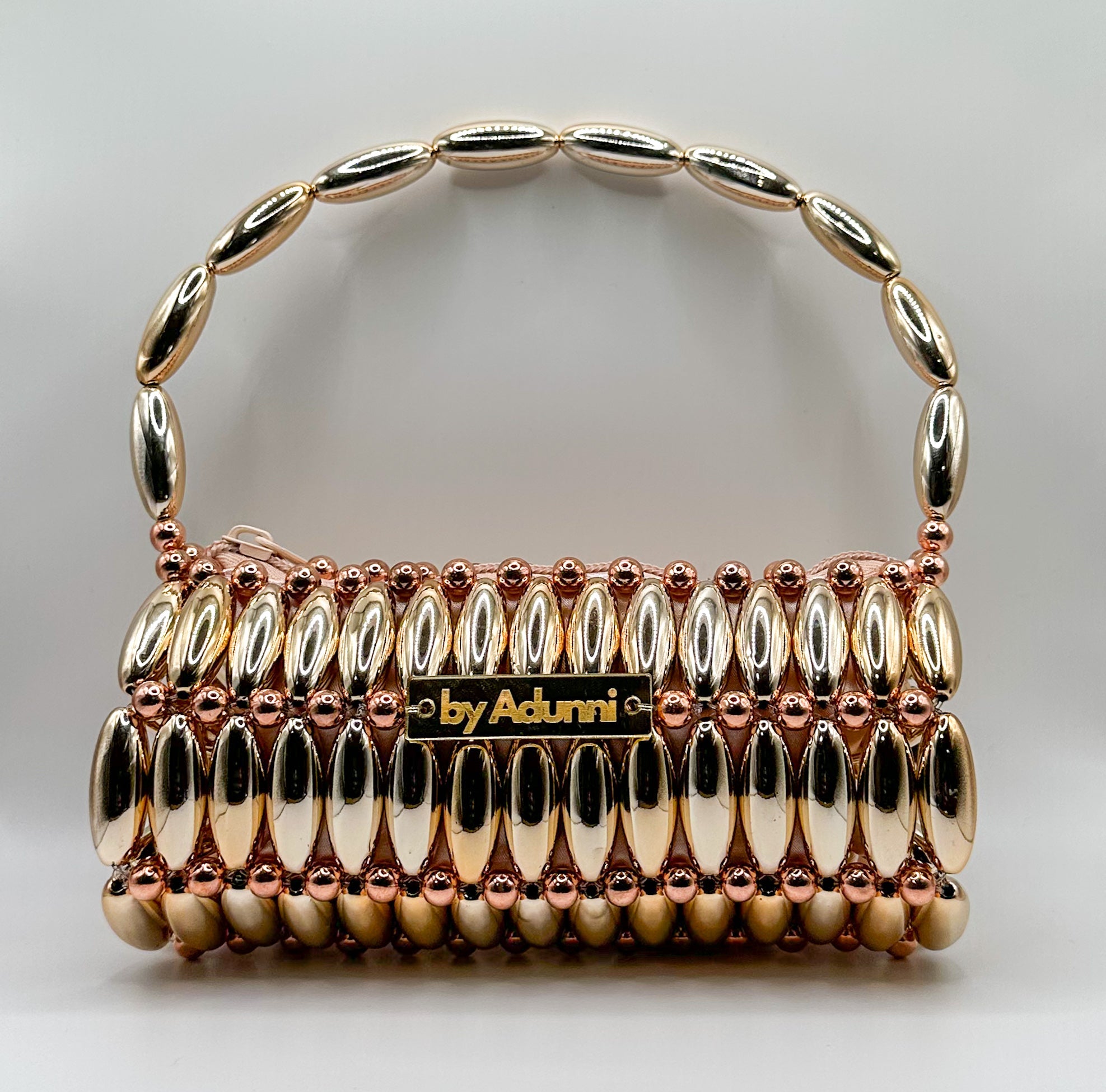 A structured handbag by Adunni, featuring a metallic gold and rose gold pebble-handle design with a central gold plaque displaying the brand name.