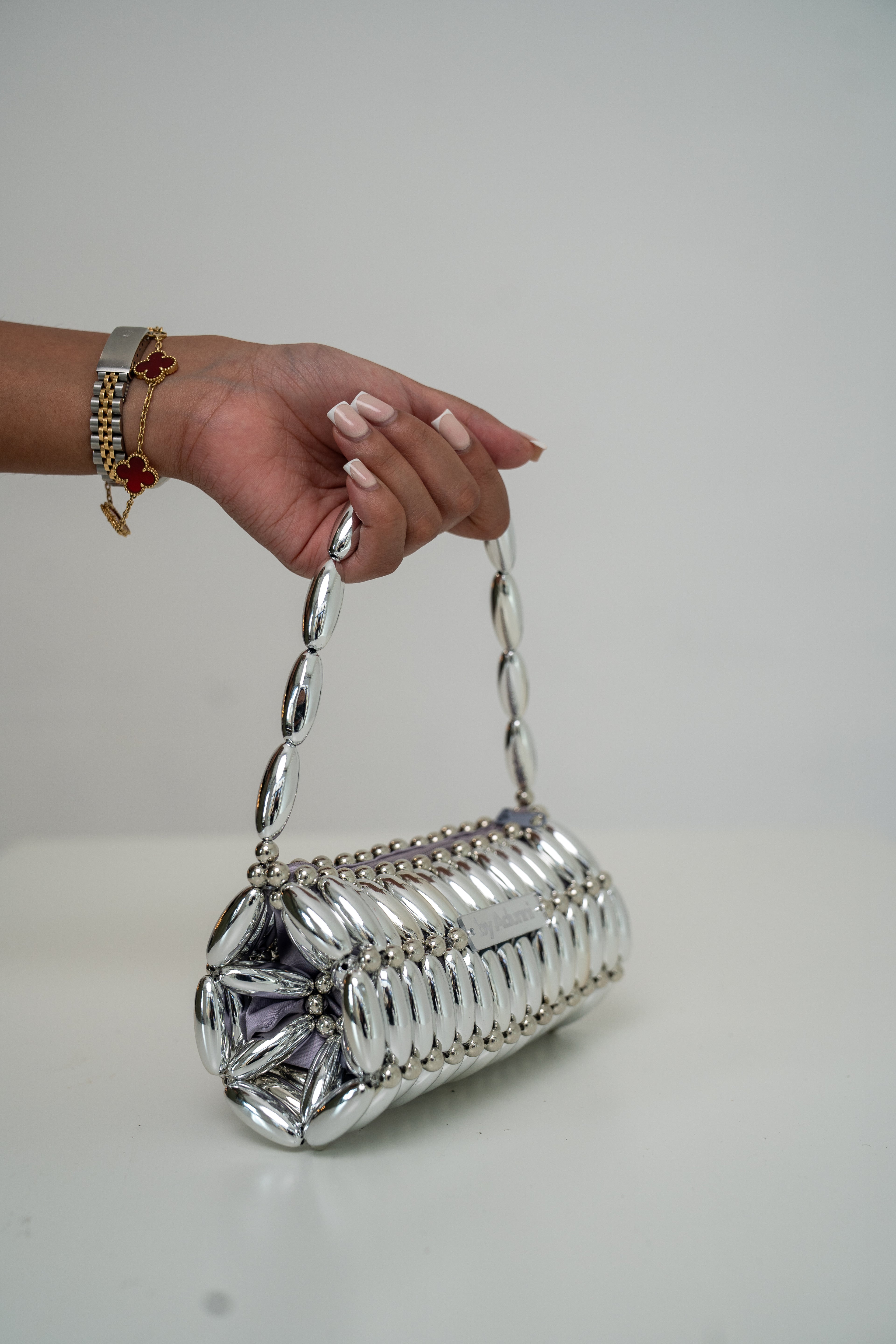 A hand with manicured nails and gold bracelets holds a metallic silver cylindrical shoulder bag with a chain strap, set against a plain light gray background.