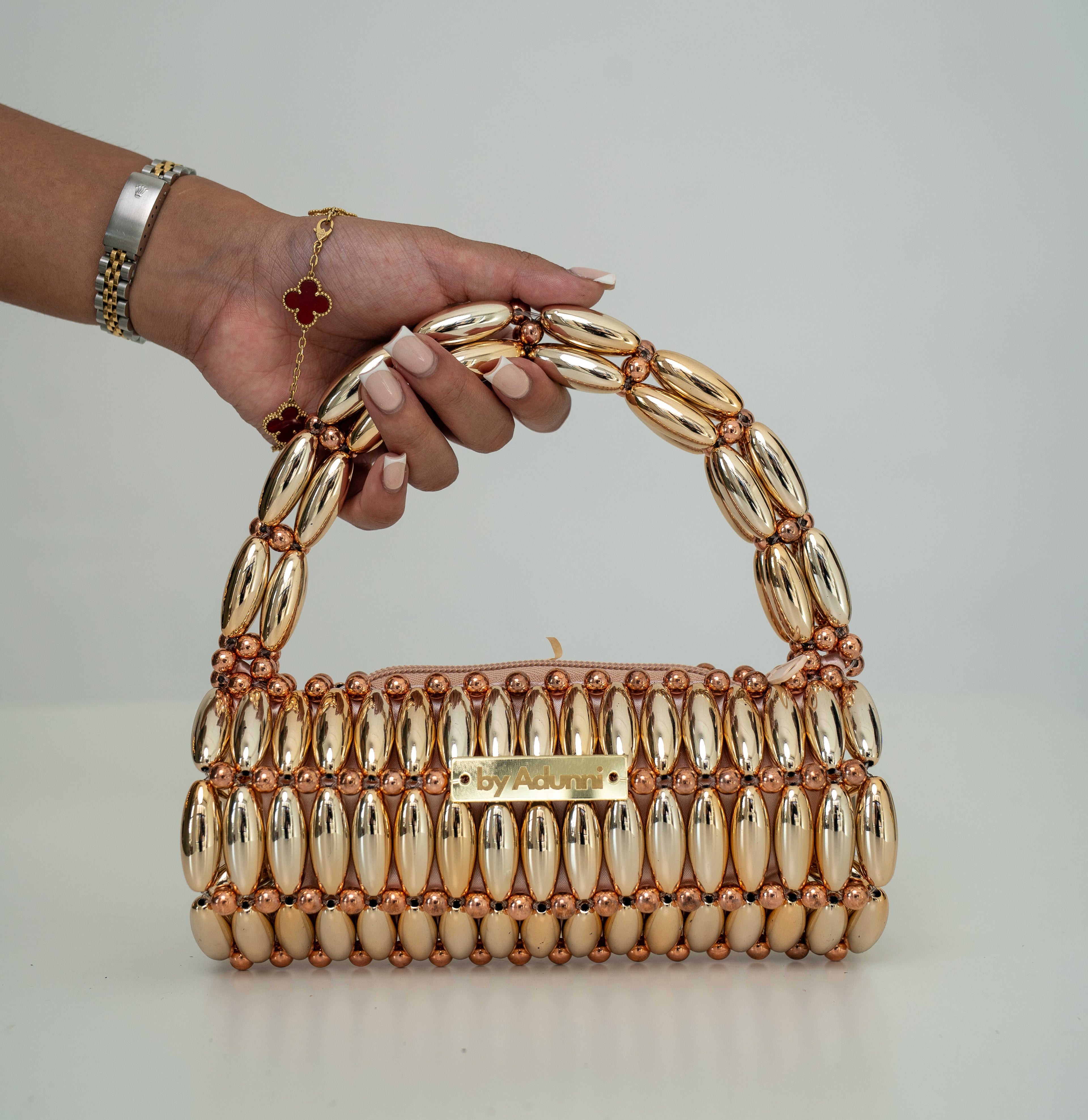 A hand holding a structured metallic gold handbag with a curved top handle, made of elongated oval links and accented with rose gold-toned beads and a small gold clasp, against a plain light gray background.