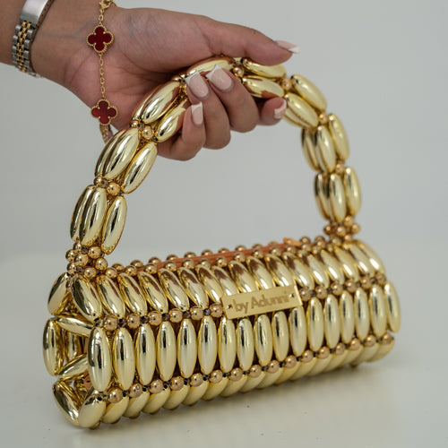 A gold metallic handbag with a cylindrical body and a matching handle, held by a person's hand. The bag features a textured, ribbed design with a small gold plaque on the front.