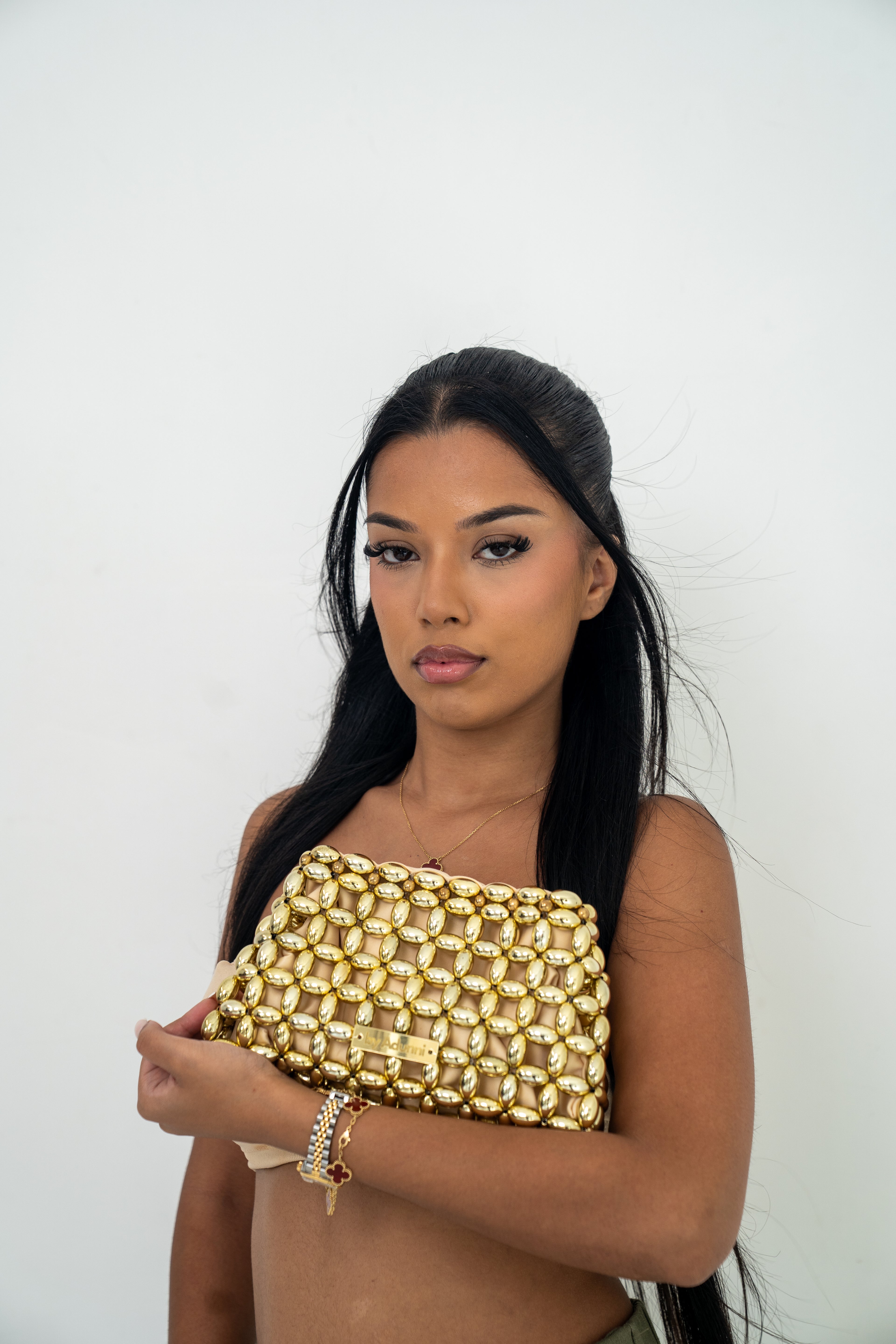 A woman with long black hair holds a gold metallic beaded clutch bag against a plain white background.