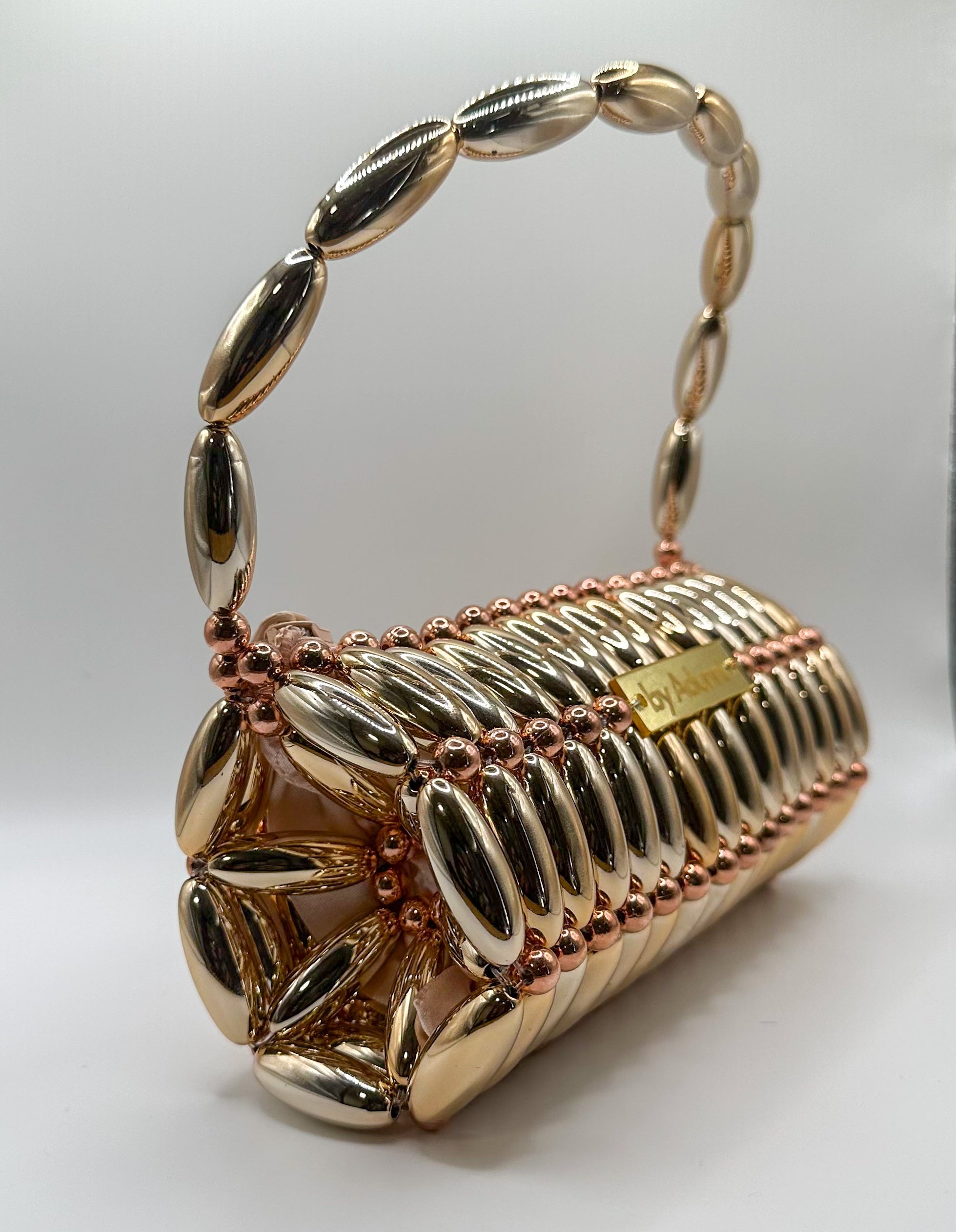 A gold metallic clutch handbag with a ribbed cylindrical body, a matching chain handle, and small rose gold bead accents, shown on a plain light gray background.